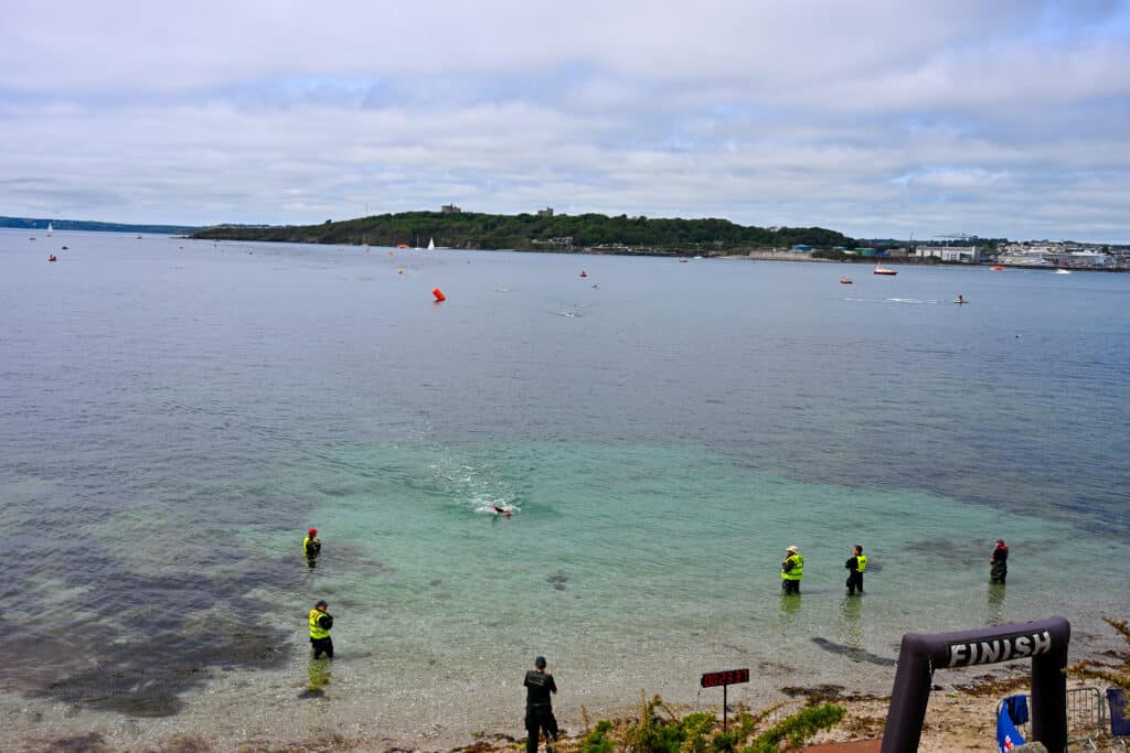 View across a body of water to a tree covered headland on the horizon. Cloudy sky above. In the foreground is a black inflatable finish line for a race. Officials in green fluorescent jackets stand in the water as a swimming approaches through the water. Looks like a swimming race.