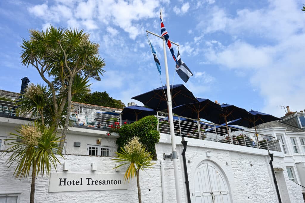 A white wall stretches across the bottom of the frame. It has a sign saying Hotel Trenton beneath a palm tree. A flagpole in the middle. Terrace with blue sun parasols above the wall. Blue sky above.