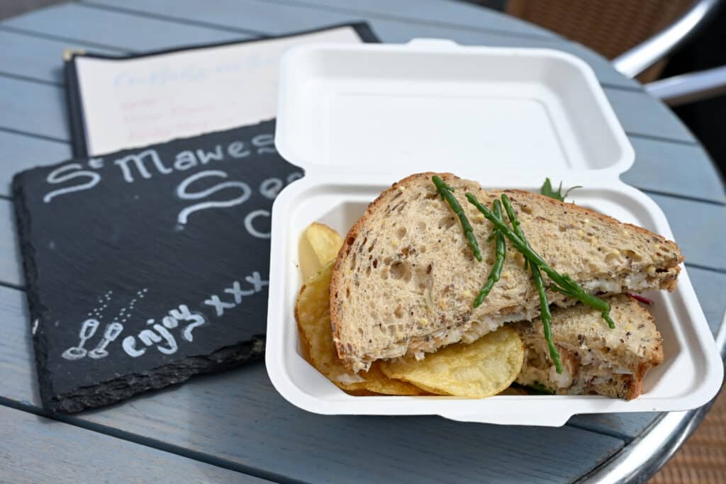 A white food tray sits open on a round grey table top. It is filled with a brown sandwich with green strands of samphire on top. Crisps underneath