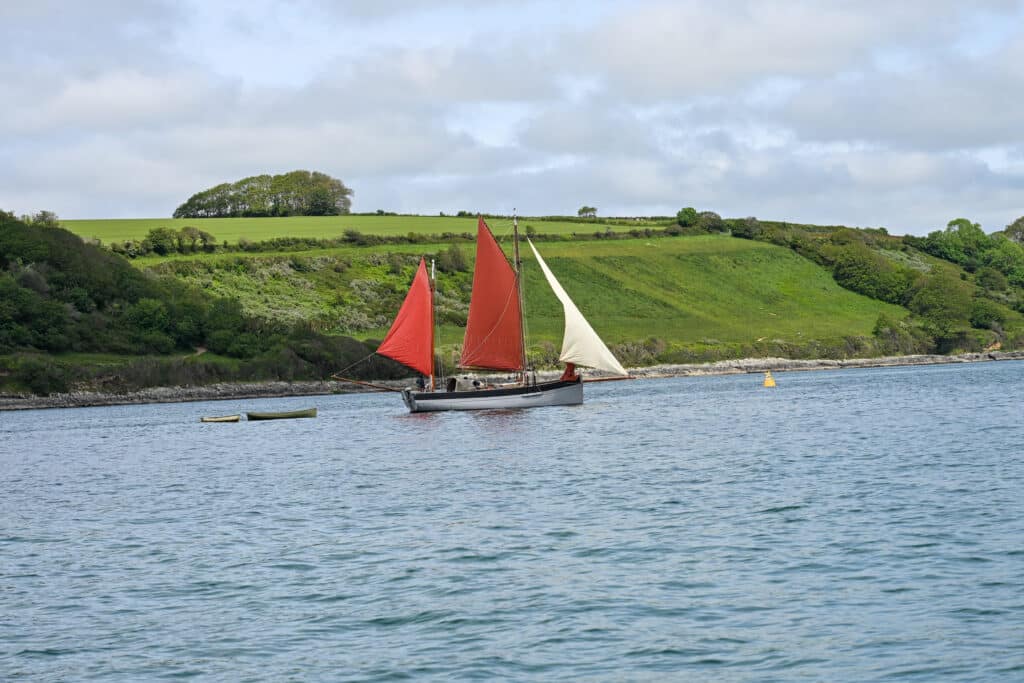 A wooden boat with three sails is moving across the middle of the frame from left to right. It has a cream sail at thhe front and two red sails in the middle and behind. It is pulling a small rowing boat at the back. Blue sky at the bottom. Green hills behind. Grey sky above.