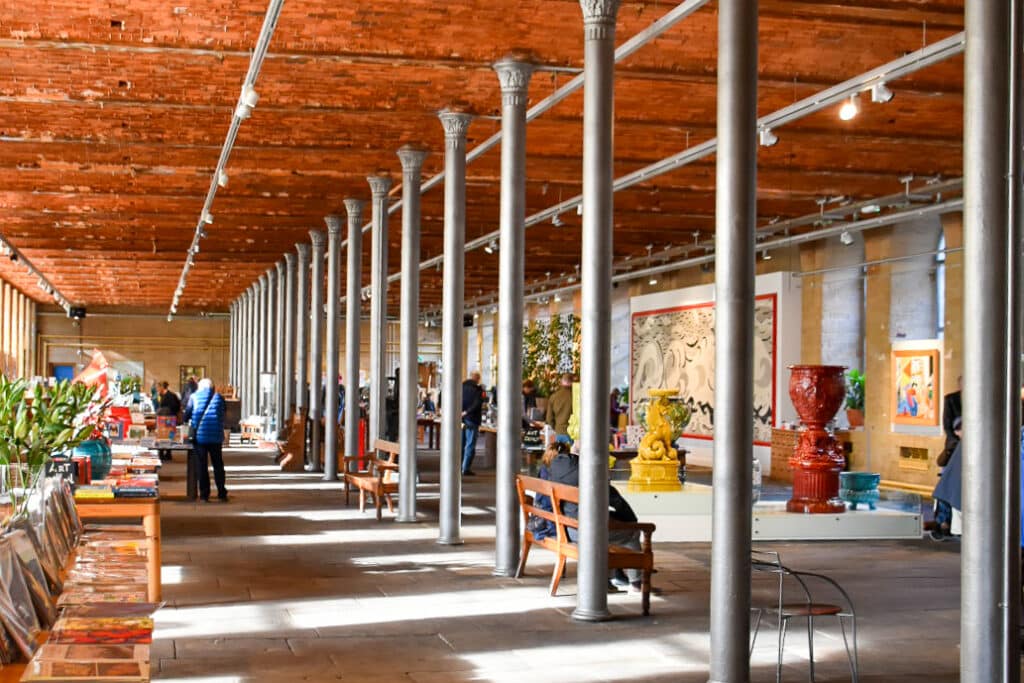 Inside a large open space with silver pillars and wooden roof. Filled with art work