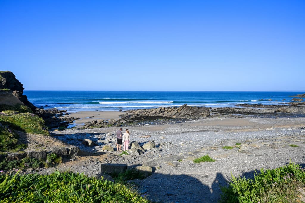 Pebbly beach with shrubs in front and sea above. two people stand on beach. Blue sky.