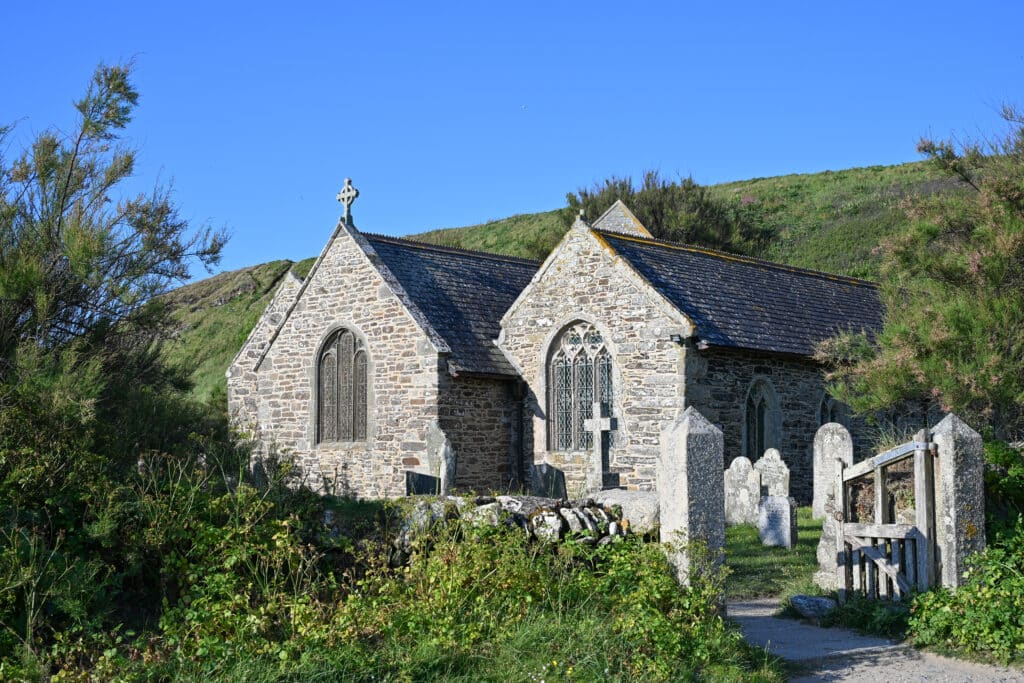 A stone church with stained glass windows with hills behind and shrubs in front. Gate and gravestones to the bottom right. Blue sky above.