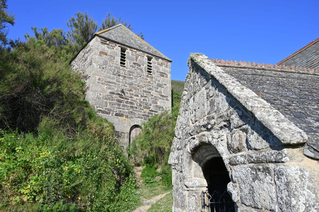 Outside a stone church entrance to the bottom right. A stone tower stands behind surrounded by shrubs. Blue sky above.