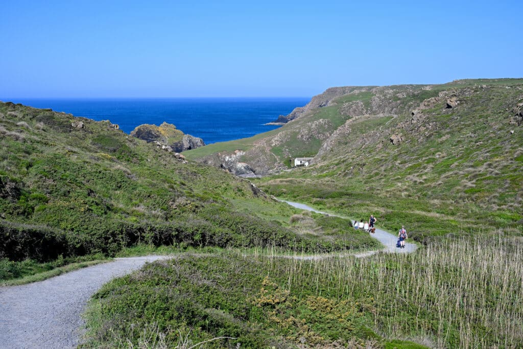 A path winds down a grass hillside towards blue water in the horizon. Pale blue sky above.