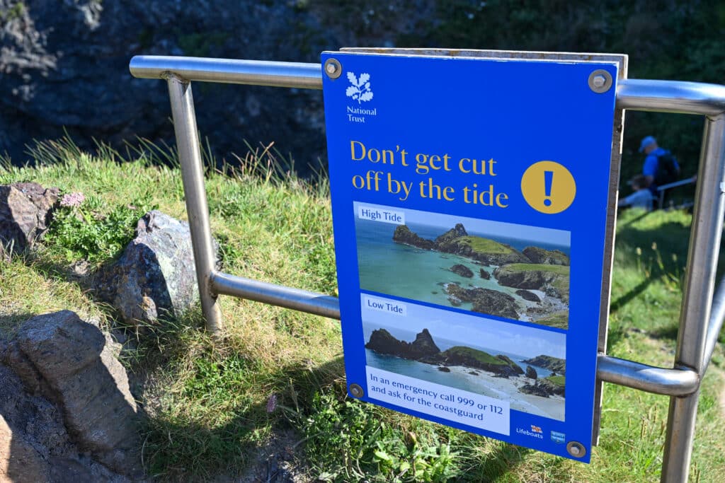 A blue sign warning you not to get cut off by the tide. Fixed to a metal frame. Grass behind.