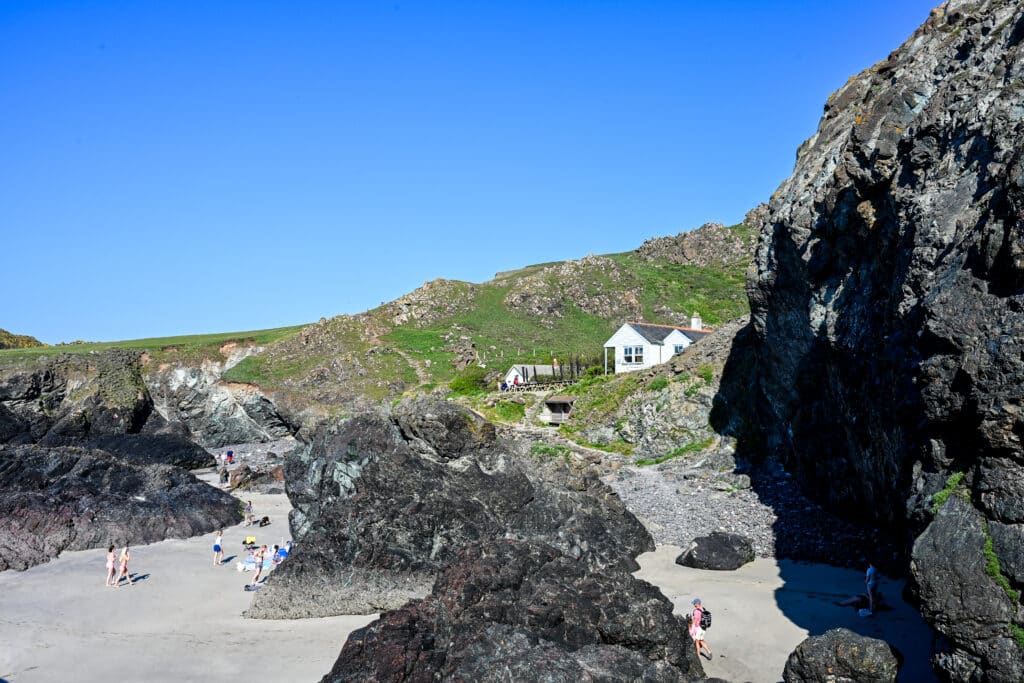 A white building sits on a rocky hillside overlooking a sandy beach with black rock stacks. Blue sky.