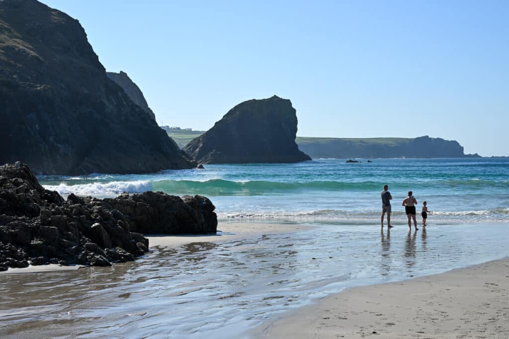 Turquoise waves rolling towards a beach around black rock stacks rising from the ground. Cliff in background. 2 adults and one child by the water