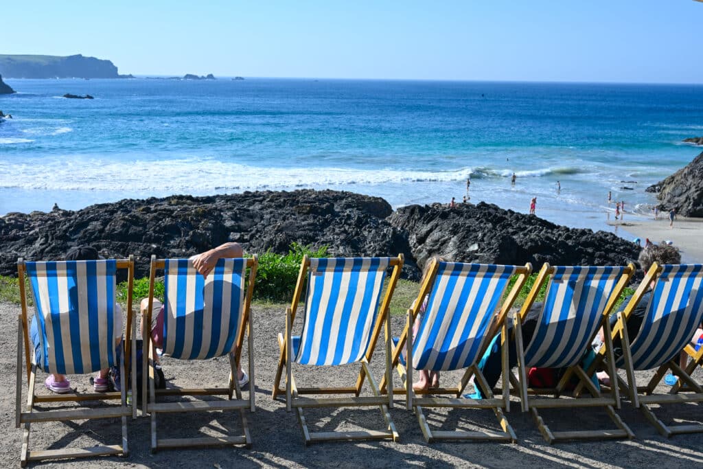 A row of blue and white deckchairs stretches across the bottom of frame, looking out at black rocks. Turquoise and blue water beyond.