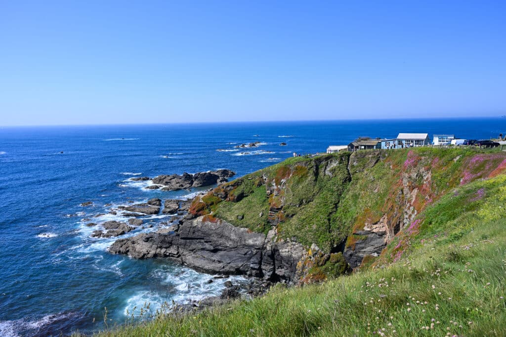 Rocky headland covered in grass. Some wooden huts on the cliff top. Blue sea and blue sky.