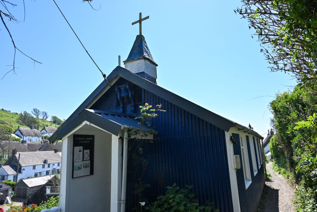 Simple wooden church stands to the left of a small path. Houses to the left. Pale blue sky above.