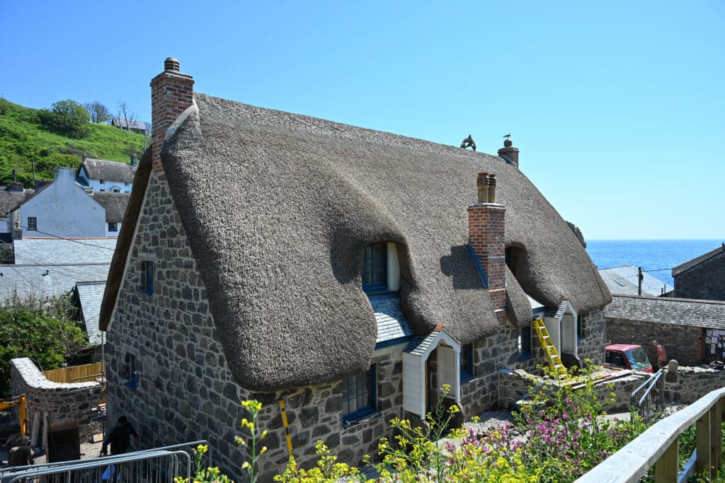 A thatched cottage stands in the middle of the frame. Other cottages behind. Sea to the right. Pale sky.