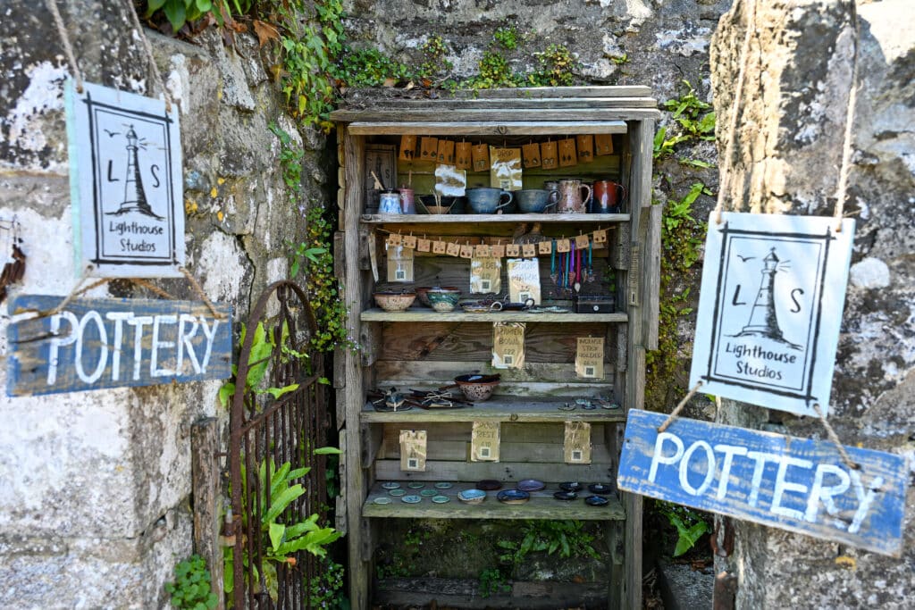 Wooden shelving unit outside against a stone wall. Filled with pottery. Signs on the side.