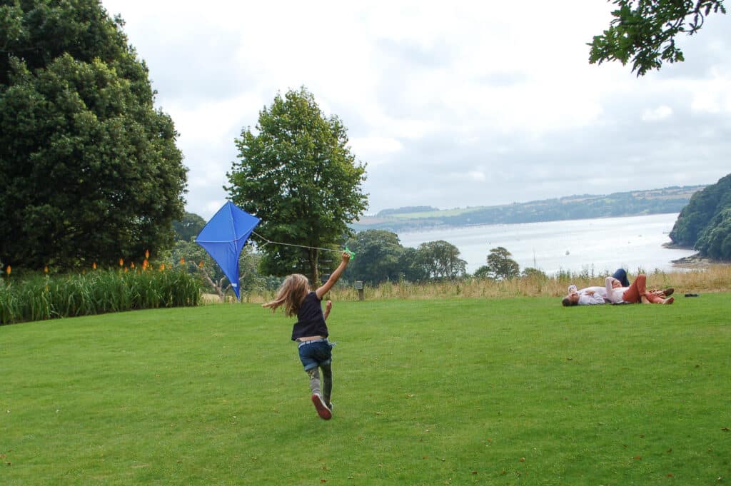 Young girl runs with a blue kite in the air, away from the camera across a lawn. Sea visible in the distance.