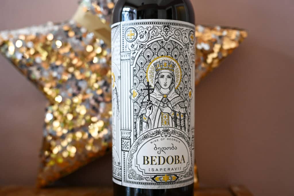 A bottle of wine in centre of photo. Pink wall and gold star behind. Beautiful drawing of female in religious robes centre of label advertising Bedoba wine.