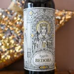 A bottle of wine in centre of photo. Pink wall and gold star behind. Beautiful drawing of female in religious robes centre of label advertising Bedoba wine.