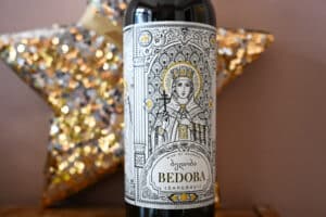 A bottle of wine in centre of photo. Pink wall and gold star behind. Beautiful drawing of female in religious robes centre of label advertising Bedoba wine.