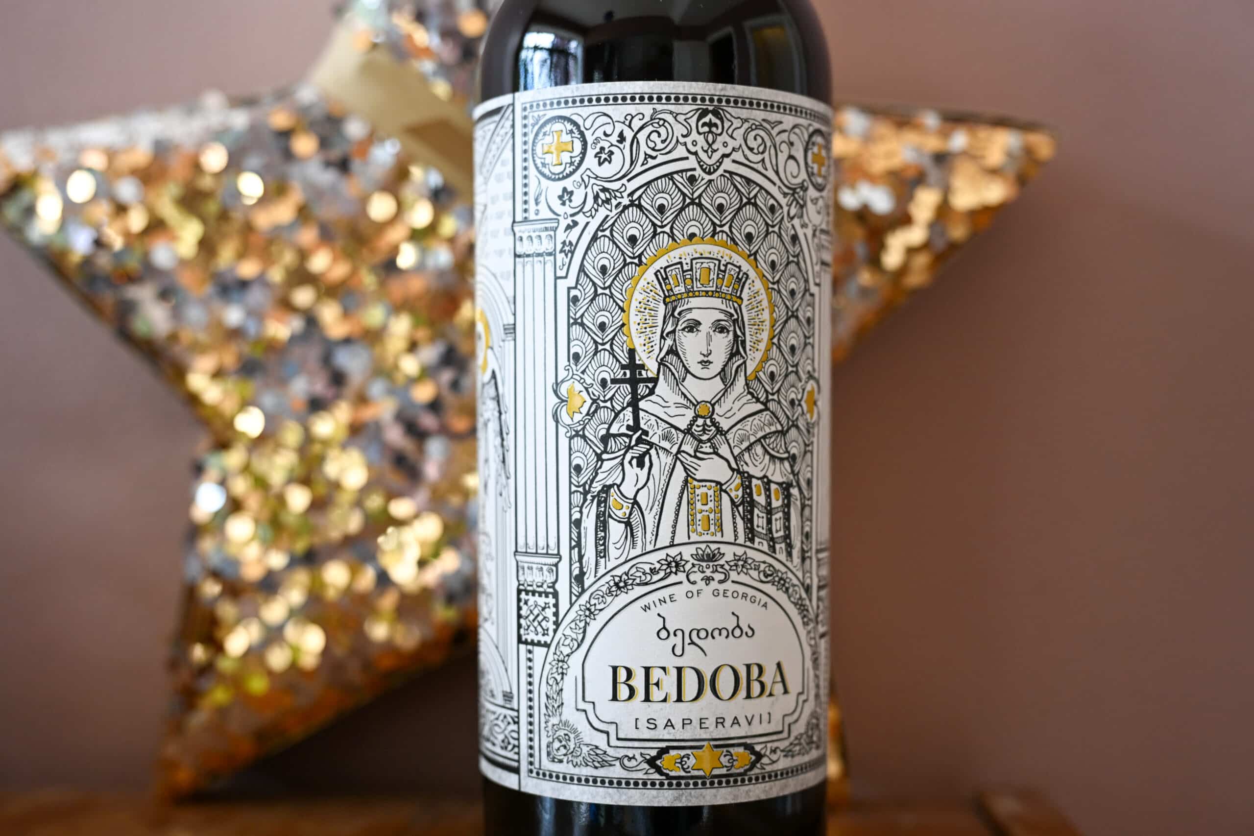 A bottle of wine in centre of photo. Pink wall and gold star behind. Beautiful drawing of female in religious robes centre of label advertising Bedoba wine.