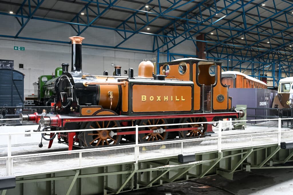 Inside a train shed. An olfactory fashioned train engine sits in the middle. It is orange with black trim. Has a funnel at the front. Says Boxhill in gold on the side.
