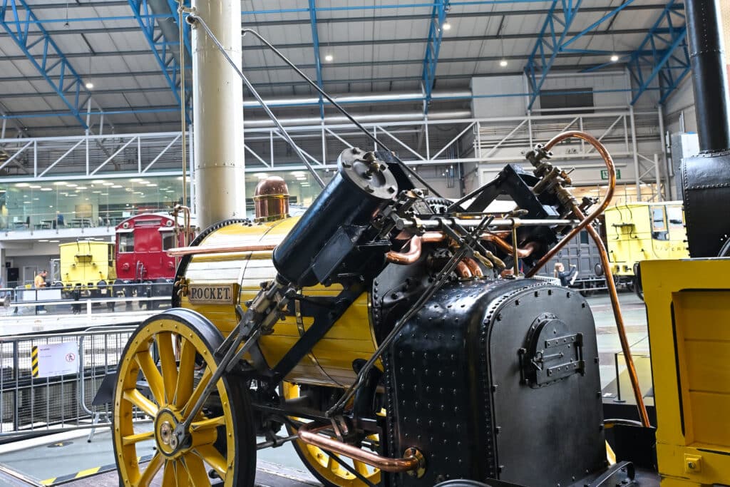 Inside a large hall. An old fashioned train sits in middle of frame. It says Rocket on the side and is painted yellow. Big wheels. Has black pistons.