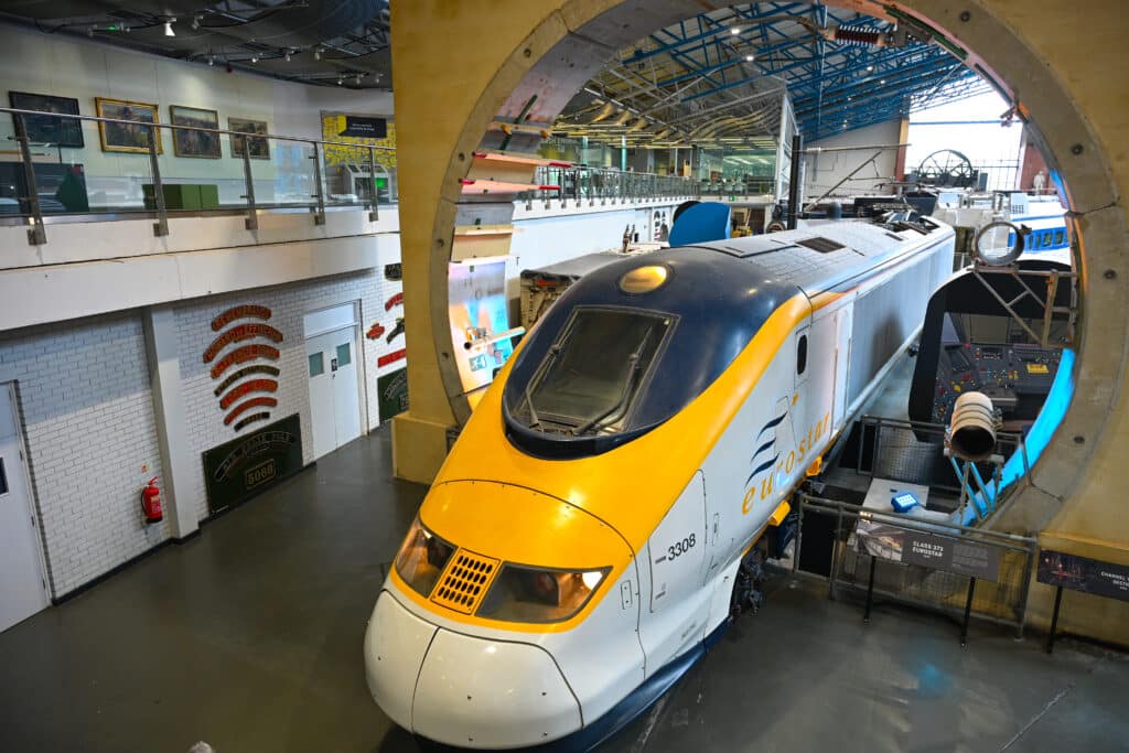 A modern train is parked facing the camera in a large hall. It has a yellow nose and says Eurostar on it. Around the engine is a circular piece of concrete.
