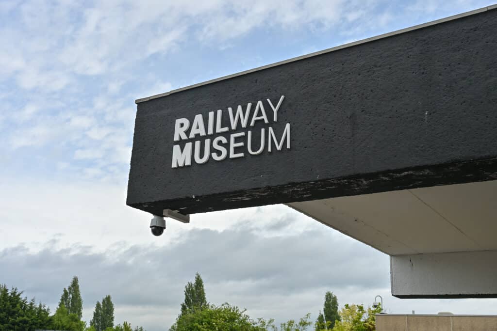 Looking up at the corner of a black roof. It says Railway Museum on it in green writing. Cloudy sky behind.