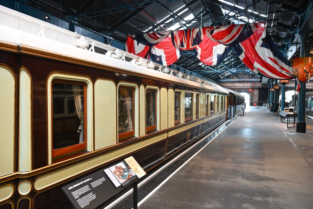 Inside train shed. Cream covered old fashioned train carriage to the left. Union Jack flags over the platform to the right.