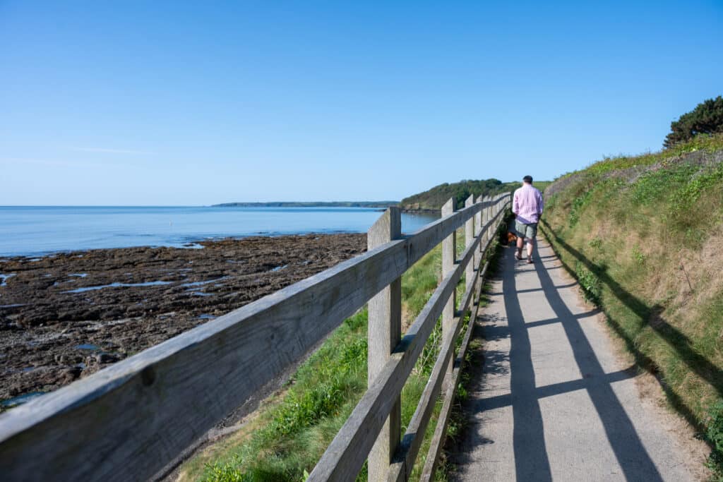 Man walks away from camera on a path. Grass to the right. Wooden fence to the left casting shadows on the path. Rocky shoreline and blue sky