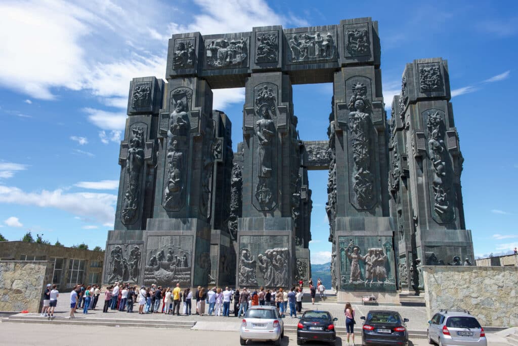 A large structure of pillars decorated with carvings of people and scenes - 5 columns across and one lying across the top. Crowd gathered in front.