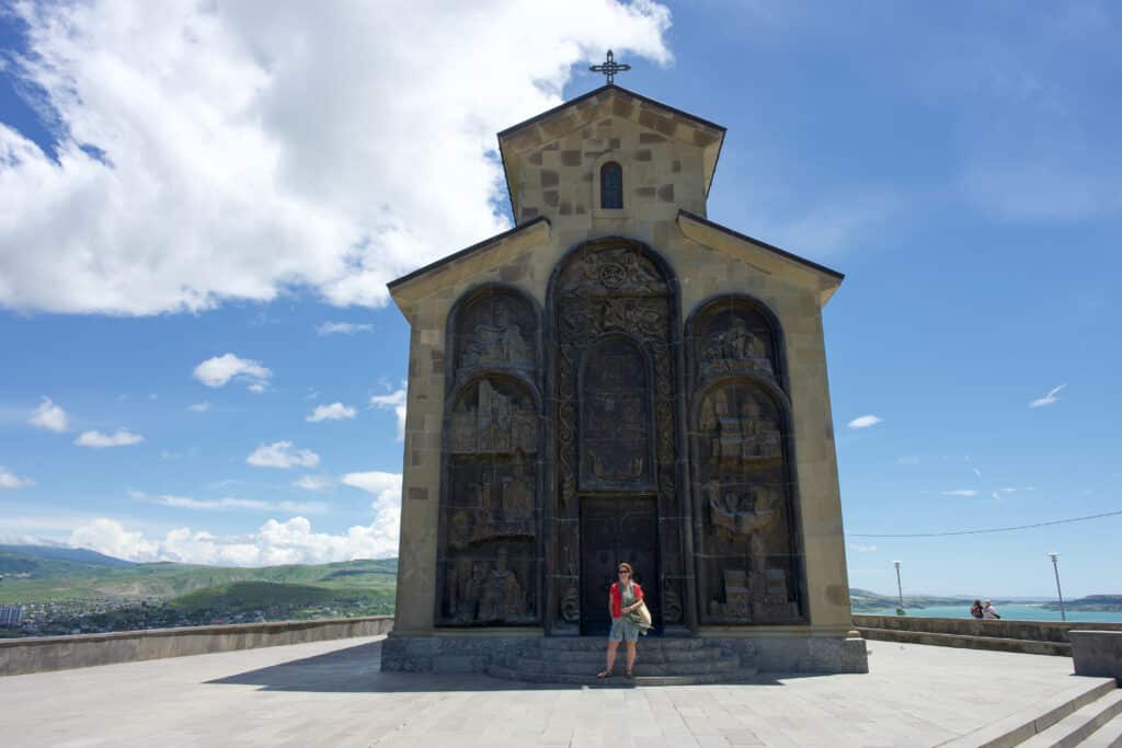 A slim church with tall wooden doors. Lady in red t-shirt in front. Pale blue sky behind.