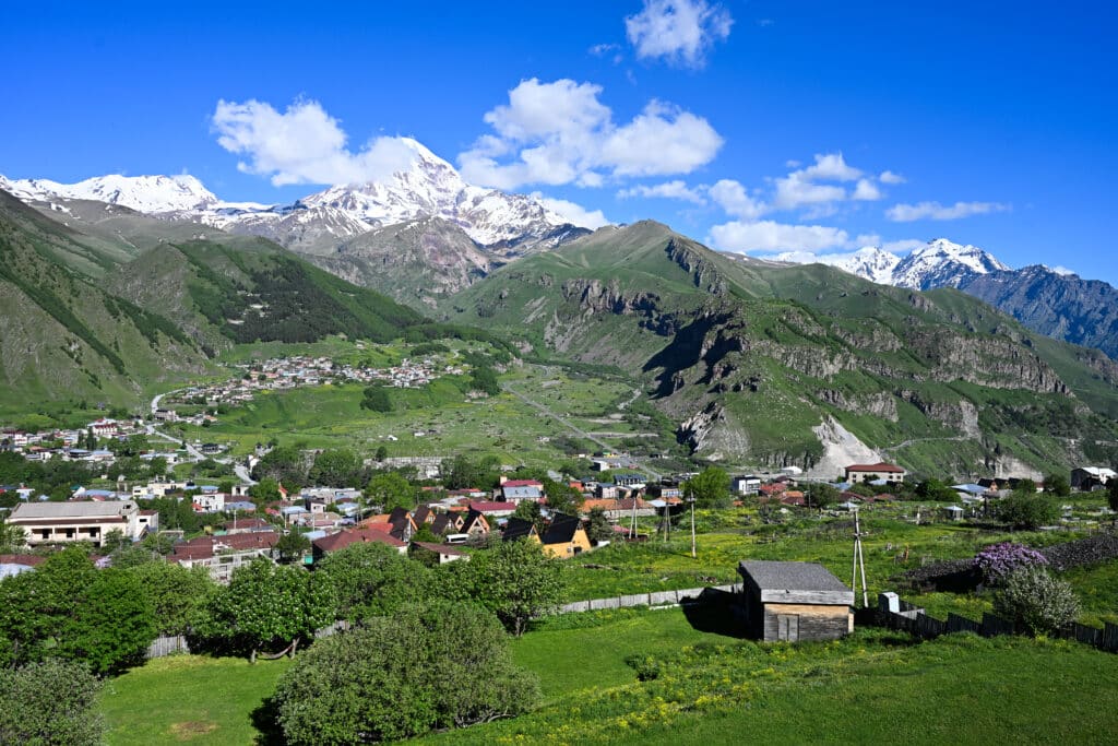 View across a green valley. Cluster of village buildings below and snow capped mountain peaks in distance.
