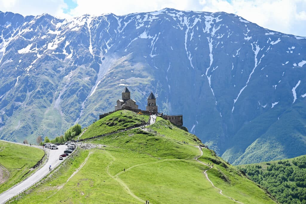 A church and bell tower stand on a green hillock with towering mountains behind
