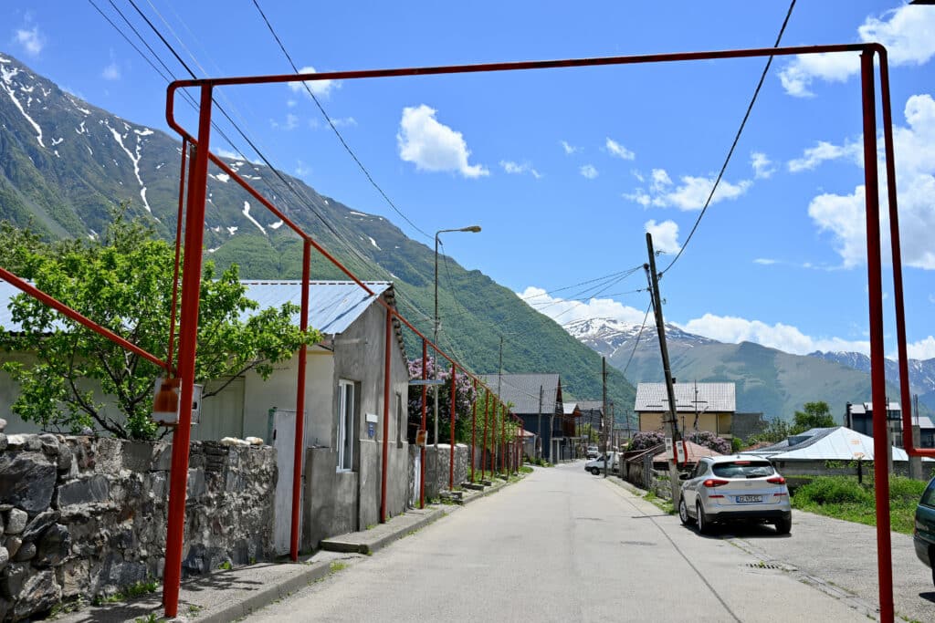 Red pipes stand like a frame over a road. Low buildings to the left. Snowy mountains behind. Blue sky