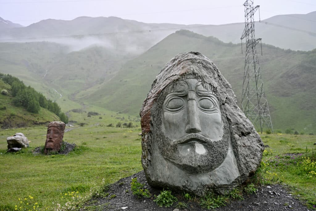 A carved male head on green-grey stone in foreground. Red carved stone head behind. Hills and mist.