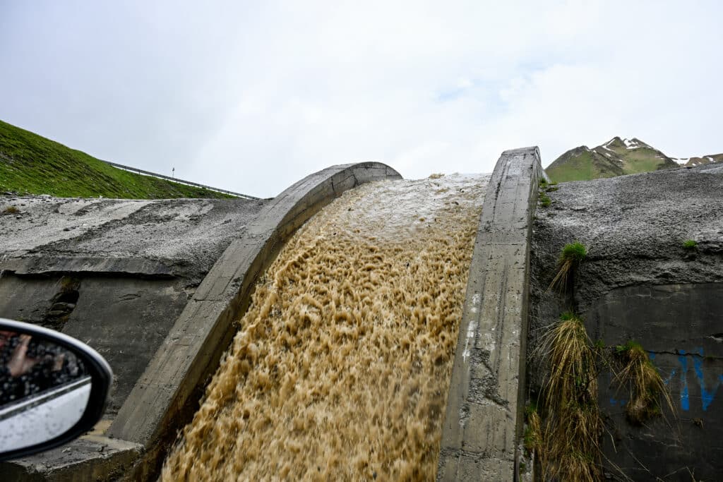 Muddy water pours over a curved stone tunnel. Mountains behind.