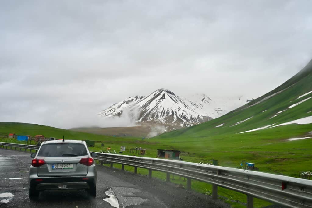 Silver car on road to the left. Green slopes to the right. Snow capped peaks in the distance. Cloud.
