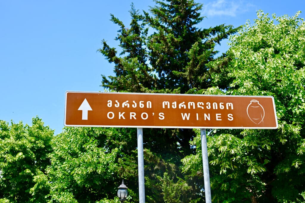 A brown road signpost sits in the middle of the frame advertising Okro's wines. Trees behind.