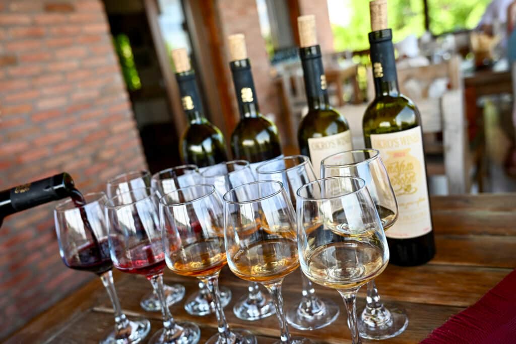 Two rows of 5 wine glasses lined up with different coloured wine samples in them. A row of wine bottles behind. Wine being poured into glass. Wine tasting.
