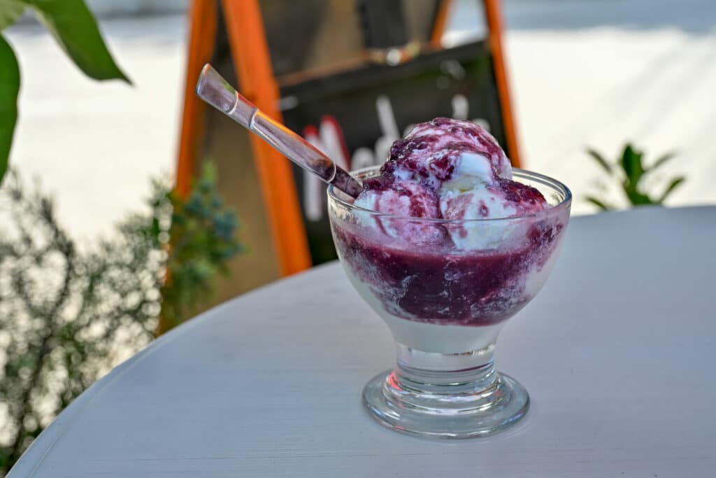 A glass bowl of white ice-cream with purple sauce and silver spoon on table