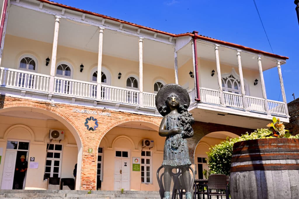 Looking up at a bronze statue of a female in dress, hat and holding flowers. A two storey building with balcony behind. Blue sky.