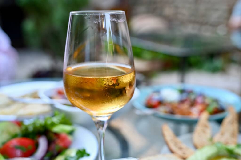 A glass of amber wine sits in the foreground. Plates of salad visible in the background (but out of focus)
