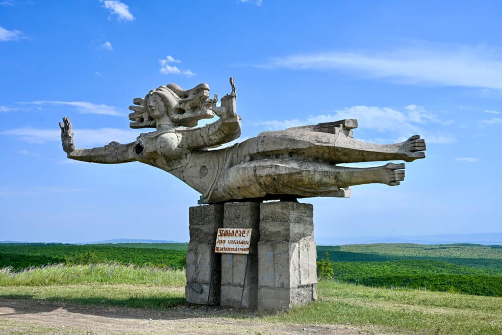 Looking at a stone sculpture of a lady lying on her side - head to the left, arms out to her sides, Looks like she's flying. Hills behind.