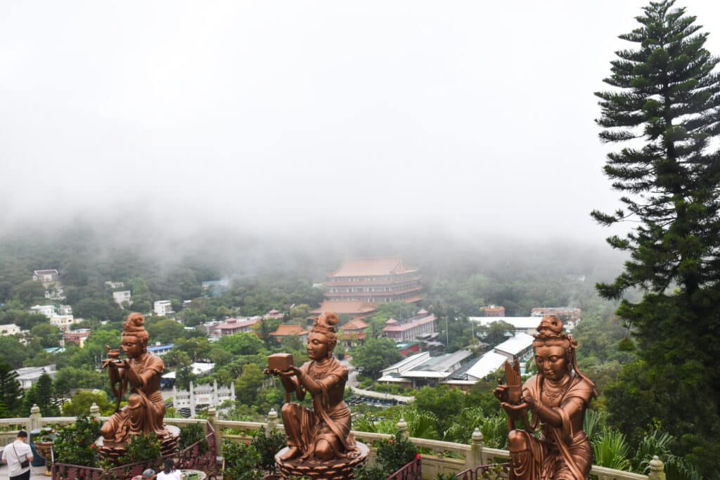 Three gold statues making offerings kneel at bottom of frame. Buildings in green valley beyond. Top half of frame filled with cloud