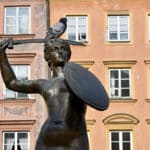 Brass statue of lady holding a shield and sword raised above head. Pigeon sitting on head. Ocre coloured building behind.