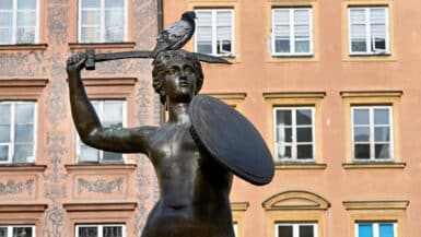 Brass statue of lady holding a shield and sword raised above head. Pigeon sitting on head. Ocre coloured building behind.
