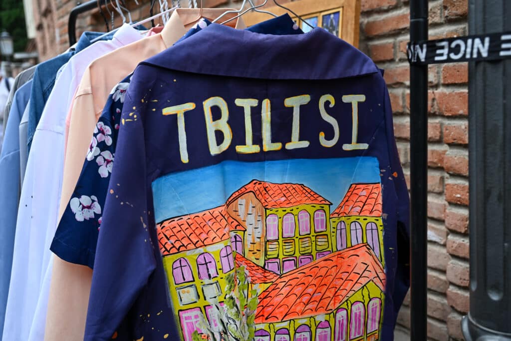 NAvy shirt with the word Tbilisi and house painted on back, hanging on a rail with other items