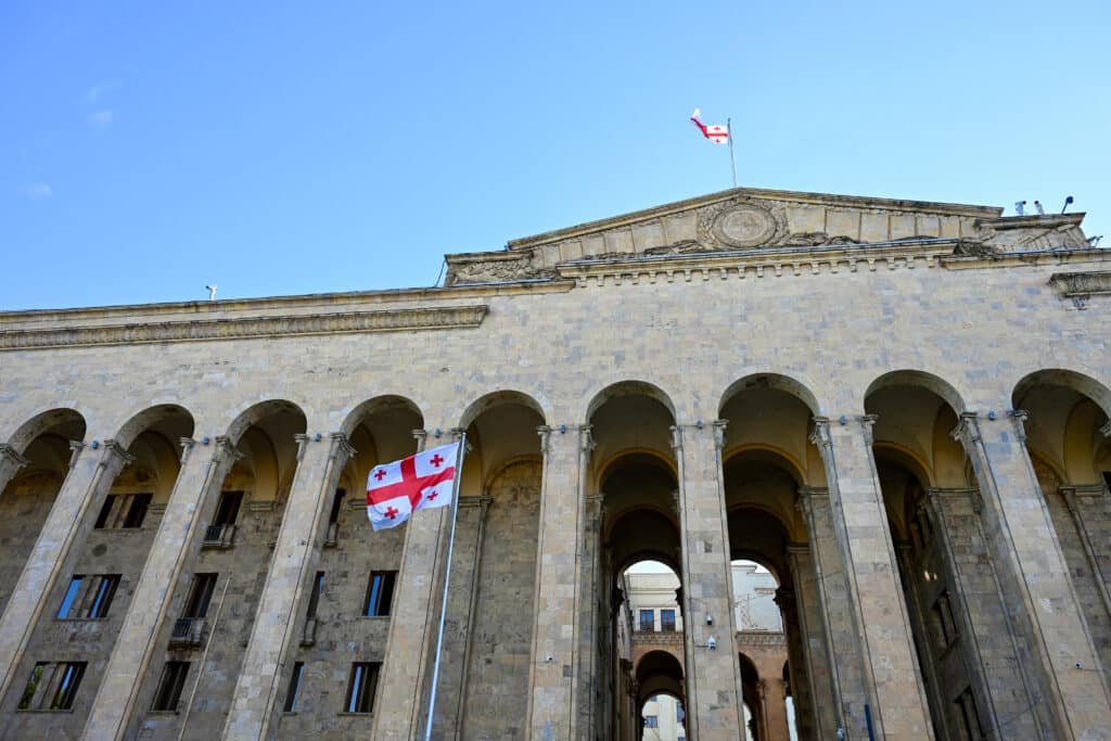 Looking up at a large stone building with arches at bottom and two white and red flags flying.