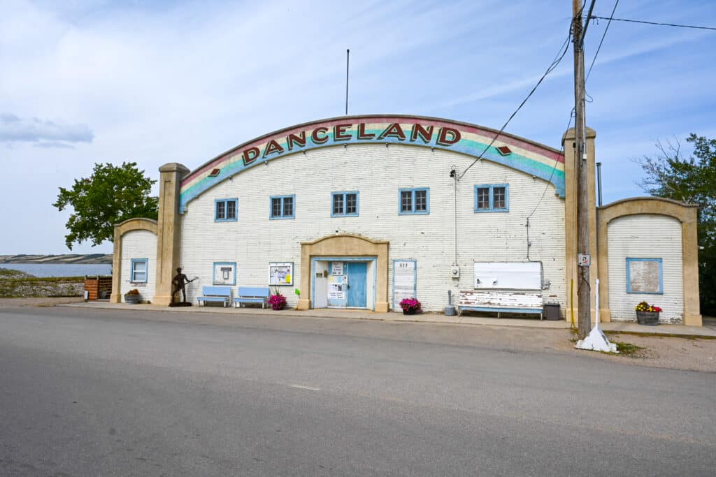 Historic Danceland dance hall in Manitou Beach, Saskatchewan — a large white arched building with a colourful rainbow “DANCELAND” sign across the curved roof, blue-trimmed windows and doors, outdoor benches, flower pots, and a guitarist statue, set beside a paved road with a lake visible in the background under a partly cloudy sky.