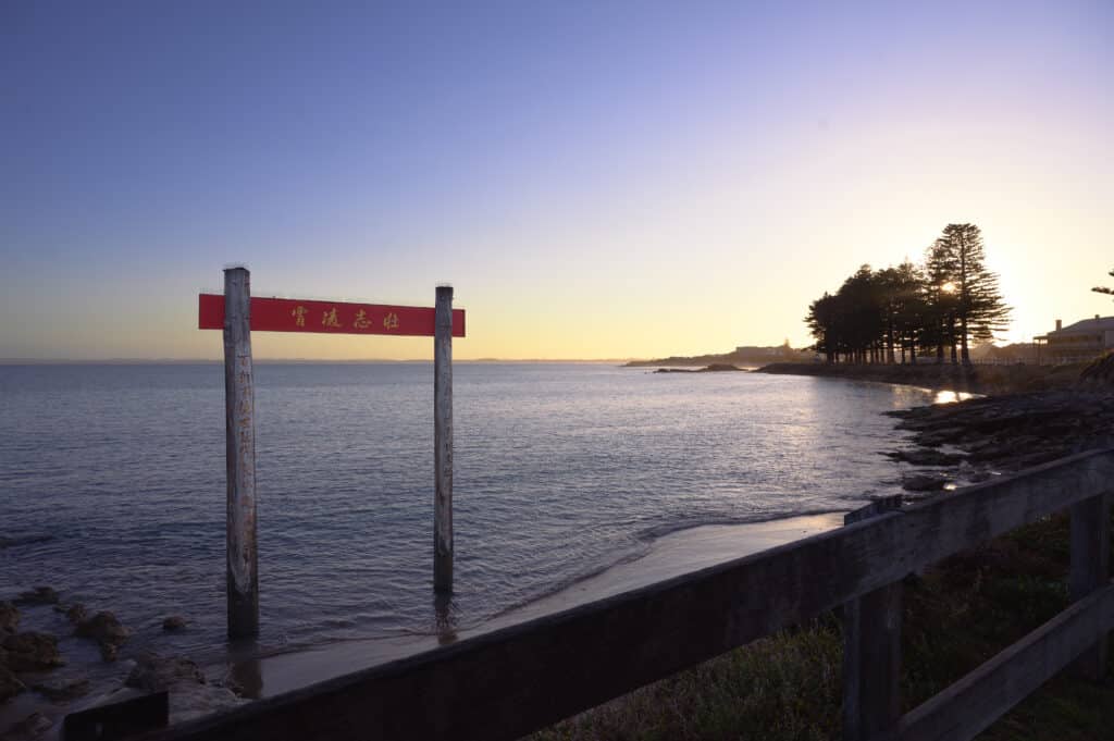 Red Chinese-style Pai Fang gate stands in shallow water at the edge of Guichen Bay, Robe, under a clear dusk sky.