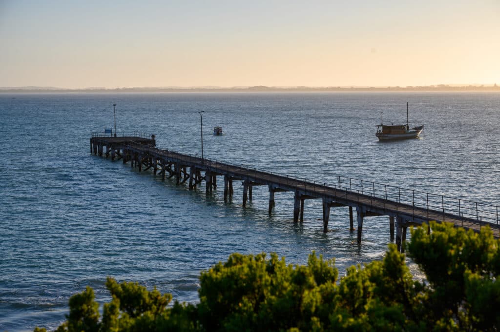 Weathered wooden jetty with railings stretches into deep blue water, two small boats visible beyond, under soft golden light.